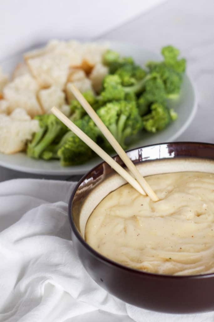 A bowl of cheese fondue on a white surface with a plate of dippers in the background.