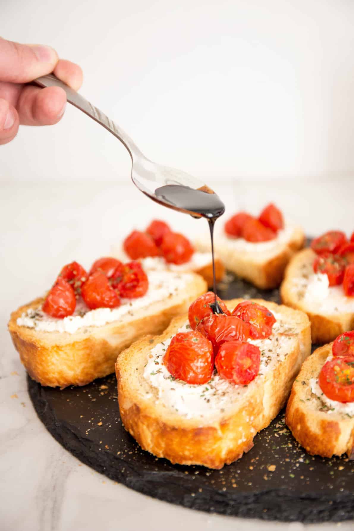 Roasted Tomato Bruschetta on a slate surface with a drizzle of balsamic reduction from a spoon.