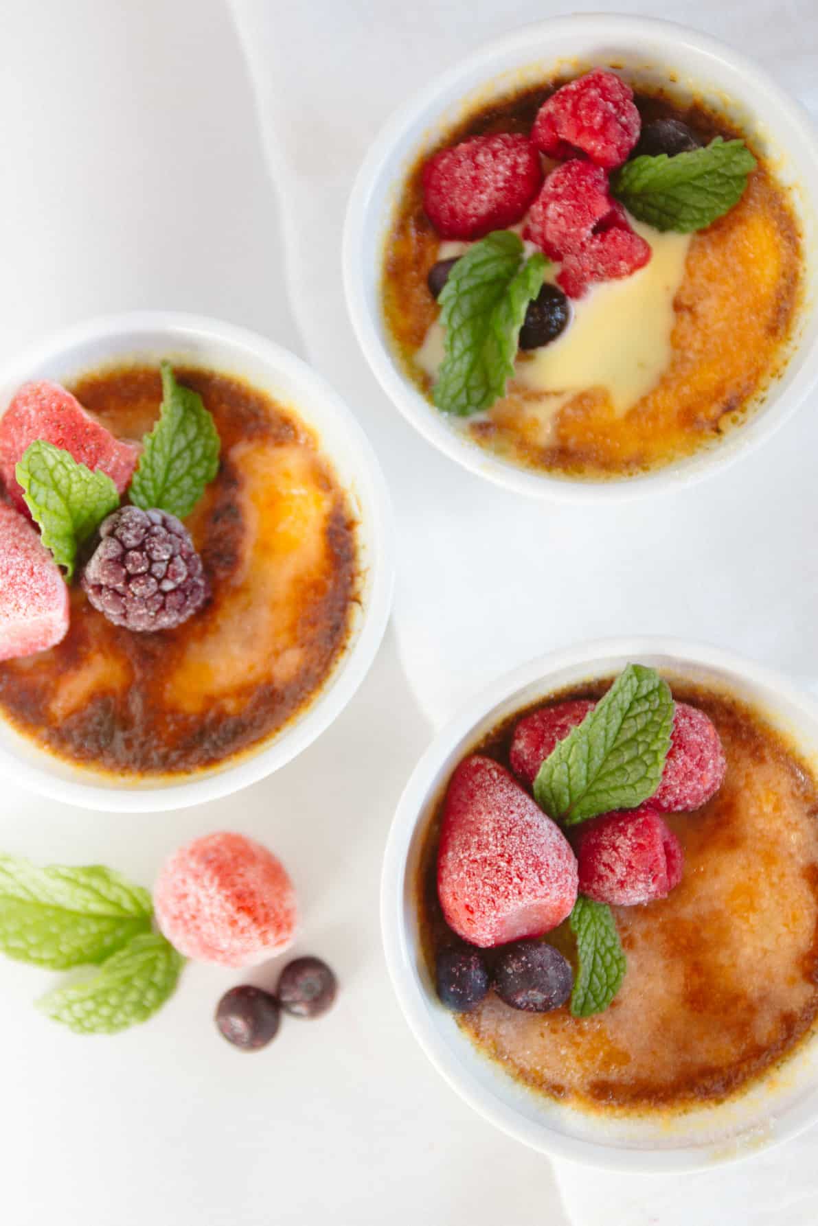 Creme brulee topped with fresh berries in white ramekins on a white surface.