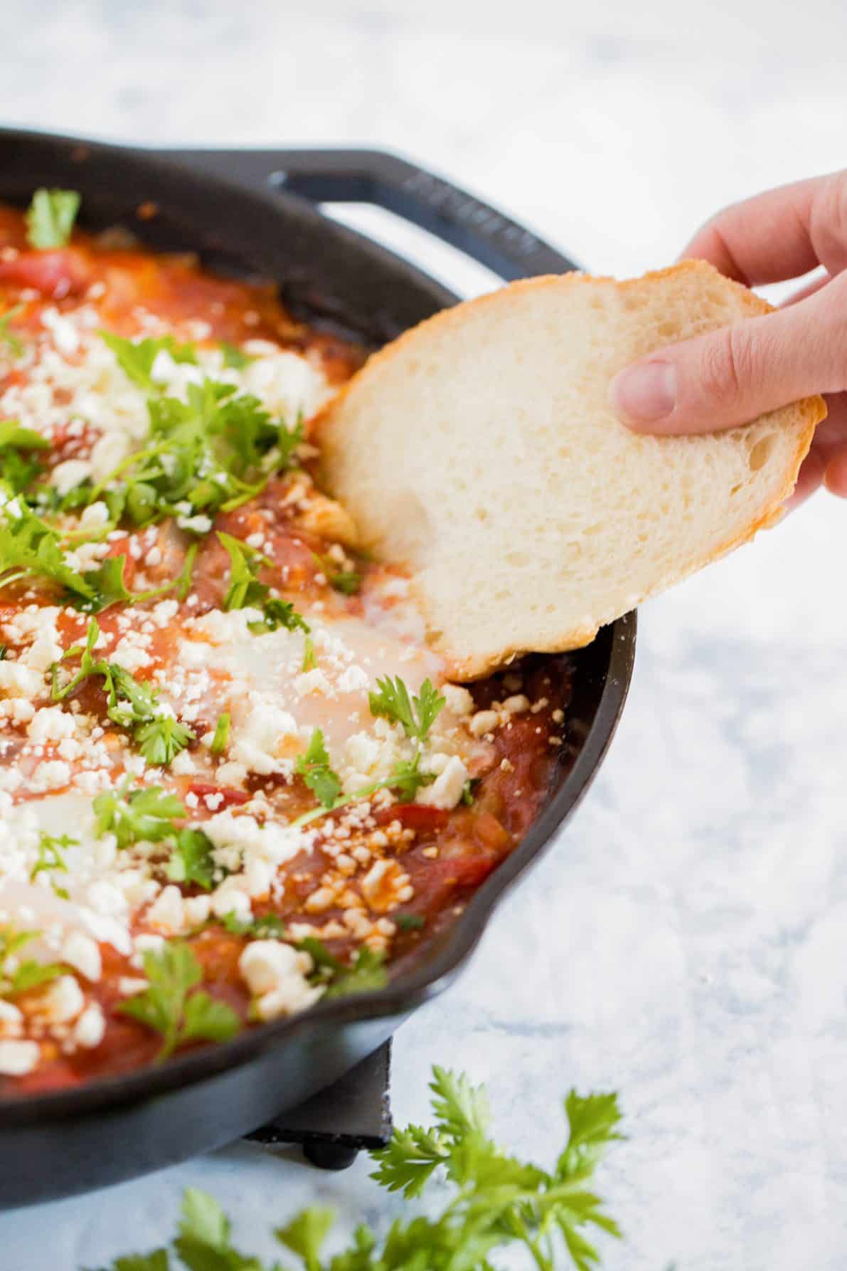 A cast iron skillet with shakshuka with feta and a hand dipping a piece of bread.