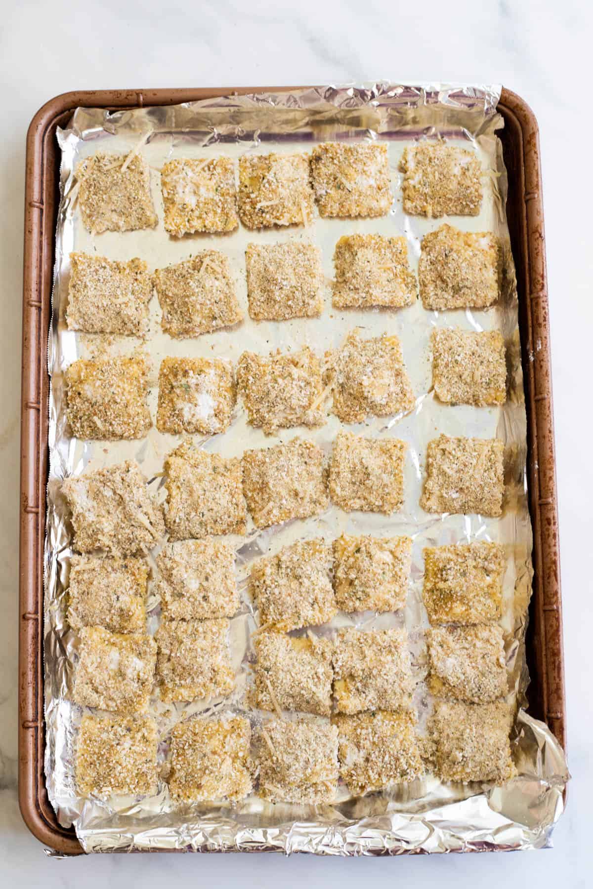Breaded ravioli lined up on a baking sheet.