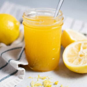 Lemon curd in a glass jar with a spoon. A sliced lemon and zest are in front.