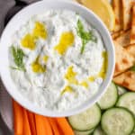 Greek tzatziki in a white bowl next to cucumbers and carrots.