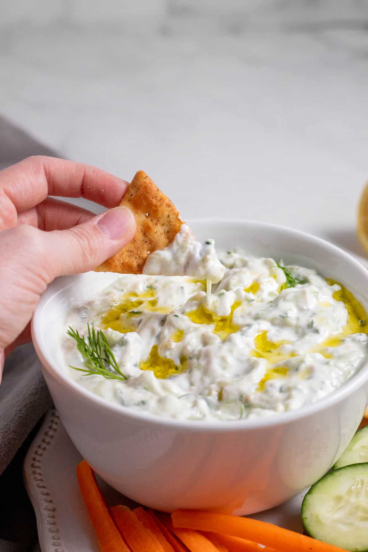 Tzatziki in a white bowl with a hand dipping a cracker into it.