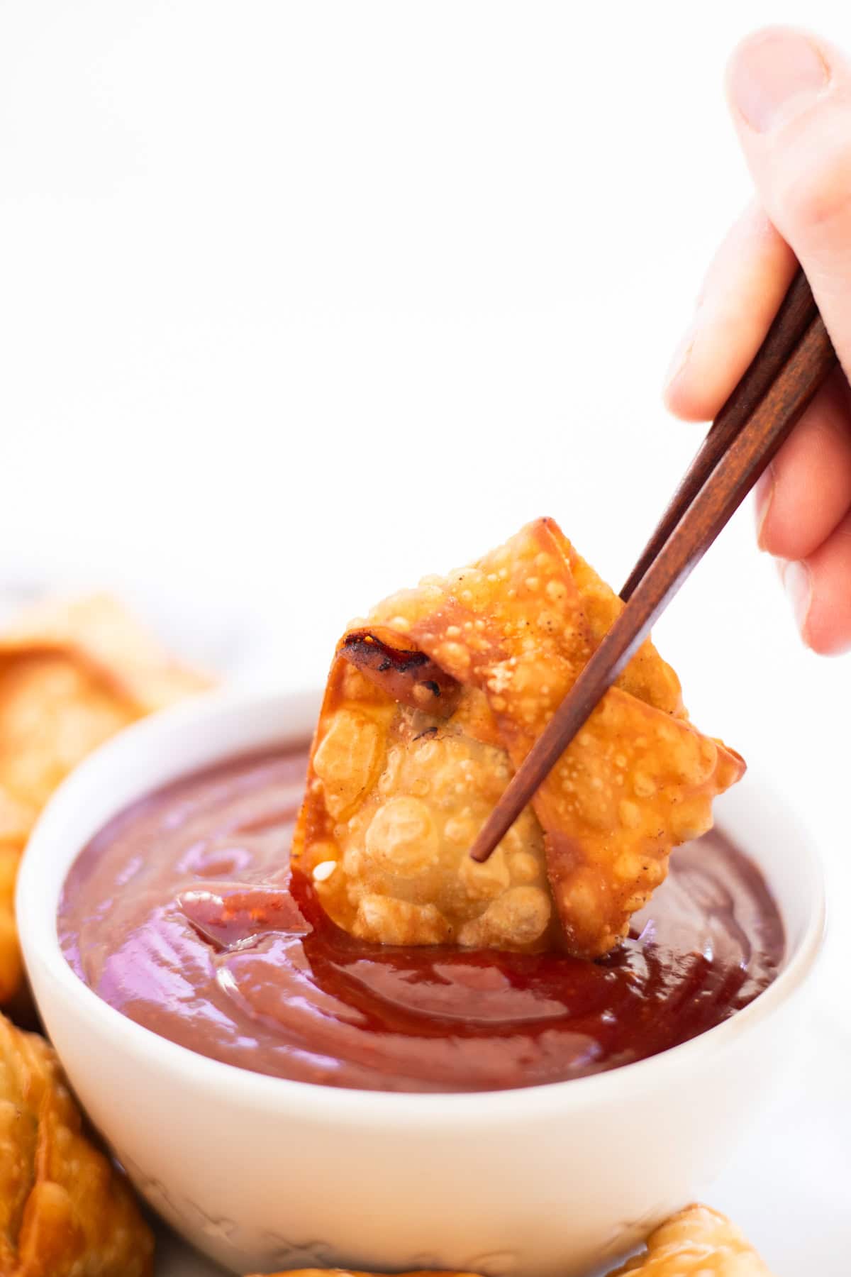A white bowl of sweet and sour sauce with chopsticks dipping a wonton into it.
