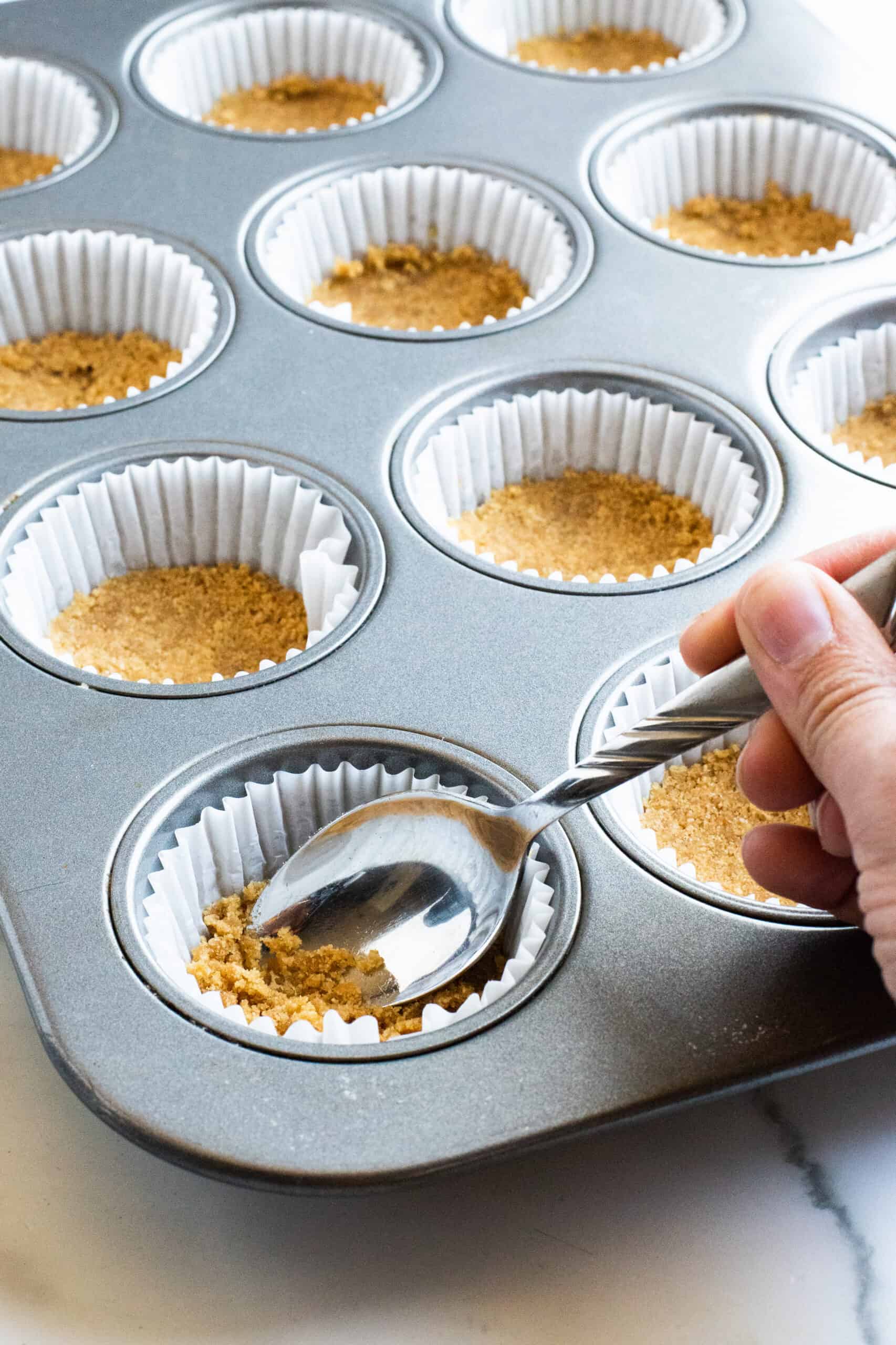 A spoon scooping graham cracker crust into the muffin tin.