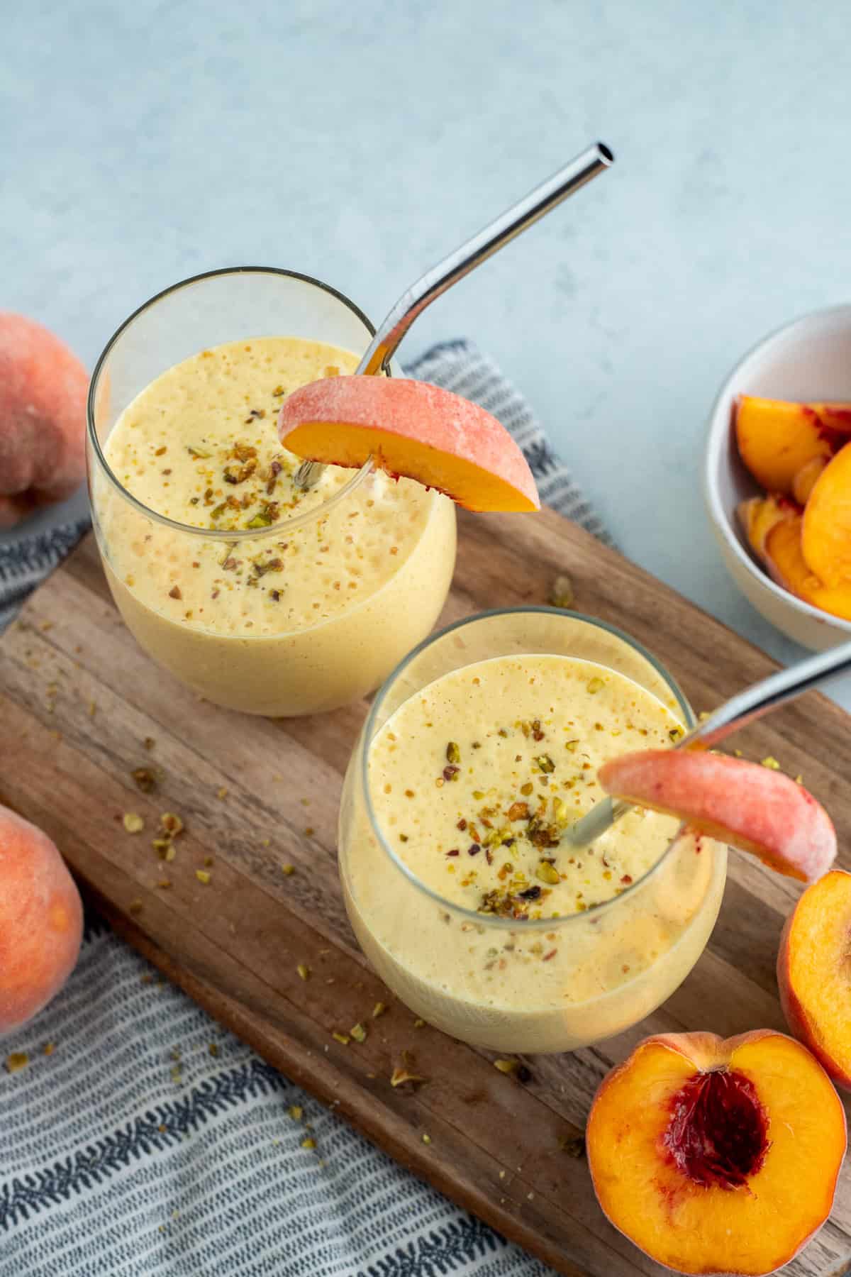 Two glasses of peach lassi garnished with metal straws and peach slices.