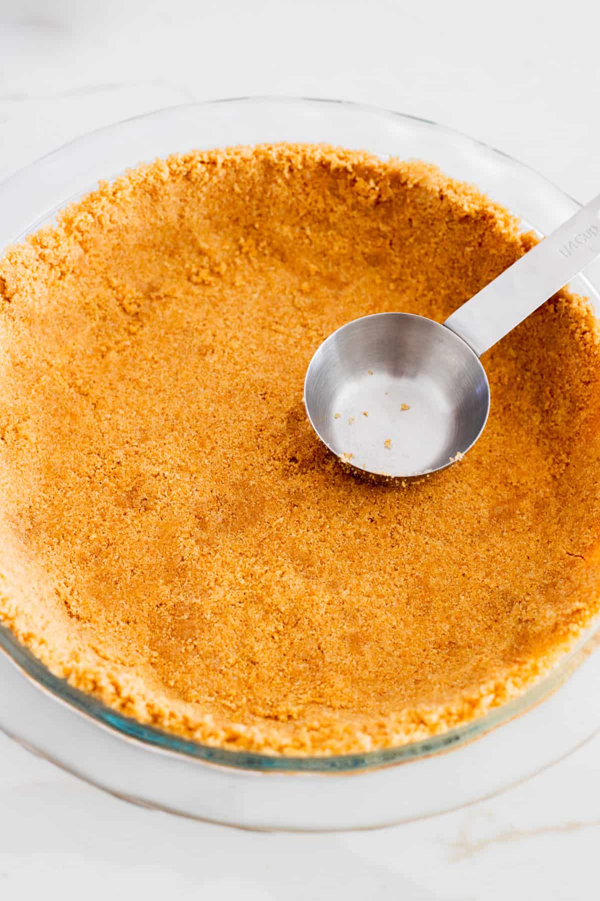 Measuring cup pressing graham cracker crust into glass pie pan.